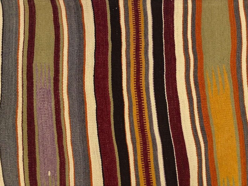 May include: A woven textile featuring a repeating pattern of vertical stripes in various colors including olive green, deep burgundy, charcoal gray, cream, mustard yellow, and black. The fabric displays a textured, flat-weave appearance.