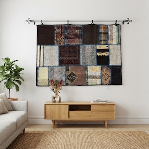 Op de afbeelding: Een rechthoekige patchwork wandhanger met diverse stoffen segmenten in bruin-, grijs-, zwart- en crèmetinten. Het textiel hangt aan een zwarte metalen roede met decoratieve krulvormige uiteinden en toont een mix van patronen en texturen.