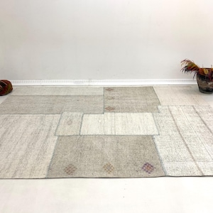 May include: A large, rectangular patchwork rug in neutral tones of beige and gray. The rug is composed of various rectangular and square patches, creating a geometric design. Decorative items, including a small cushion and a basket with dried plants, are in the background.