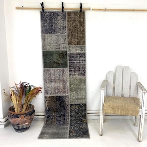 May include: A patchwork runner rug with a distressed, vintage appearance. The rug features a variety of muted colors, including greens, browns, and grays, arranged in rectangular patches. The rug is hanging on a wooden rod against a white wall.