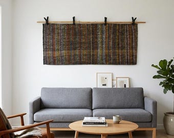 May include: A living room scene featuring a gray sofa, a wooden armchair, and a round coffee table. Above the sofa hangs a rectangular woven wall hanging with vertical stripes in brown, gold, and green. A potted plant is on the right.