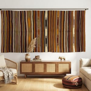 May include: A large rectangular wall hanging featuring vertical stripes in shades of brown, tan, cream, black, and deep red. The textile is suspended from a dark metal rod mounted horizontally against a plain white wall above a wooden console.