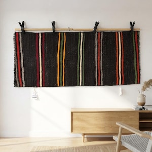 May include: A rectangular black woven wall hanging featuring vertical stripes in red, yellow, green, and white. The textile is suspended from a horizontal wooden rod by four black clips. The fabric has a textured appearance with fringed edges.