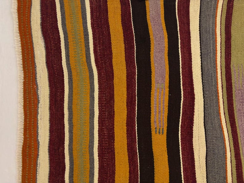 May include: A woven textile featuring vertical stripes in shades of deep burgundy, mustard yellow, black, cream, olive green, and soft lavender. The fabric has a visible textured weave and a fringed edge along the left side with a small embroidered detail.