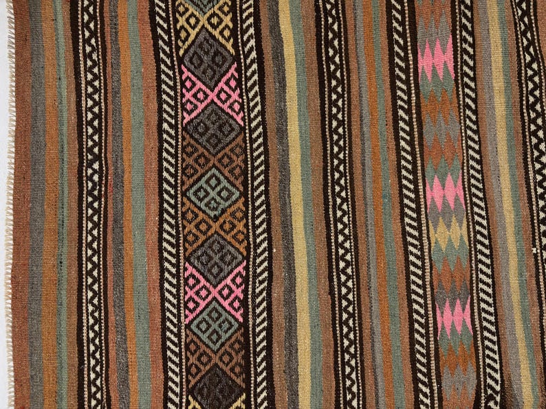 Op de afbeelding: Een geweven textielkleed met verticale strepen in tinten bruin, beige, grijs en gedempt groen. Het ontwerp bevat herhalende geometrische ruitpatronen in roze, donkergrijs en bruin, geaccentueerd door smalle zwart-witte zigzagranden.