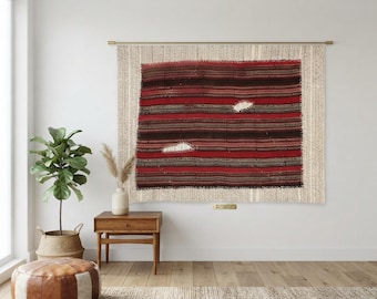 Puede incluir: Un tapiz textil rectangular tejido con rayas horizontales en tonos rojo intenso, marrón oscuro y negro. La pieza presenta dos parches blancos distintos, texturizados y con forma de nube integrados en el patrón de rayas.