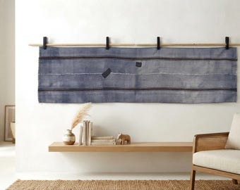 May include: A long, rectangular blue textile wall hanging featuring horizontal dark stripes and two small, dark rectangular patches. The fabric is suspended from a wooden rod by four black clips attached to the wall.