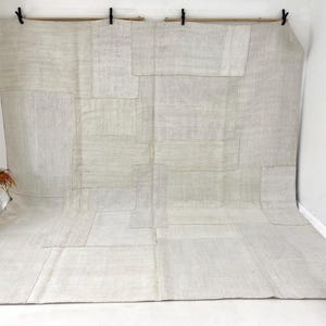 May include: A large, neutral-toned rug with a patchwork design. The rug is made of rectangular and square pieces of fabric sewn together. The colour is a light beige, with variations in tone. The rug is displayed against a white wall, with a decorative pot and dried plants in the corner.