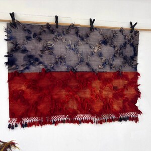 May include: A rectangular textile art piece, divided horizontally into two sections. The top half is a muted blue-gray with textured, raised elements. The bottom half is a vibrant red with a fringed edge, creating a striking contrast.