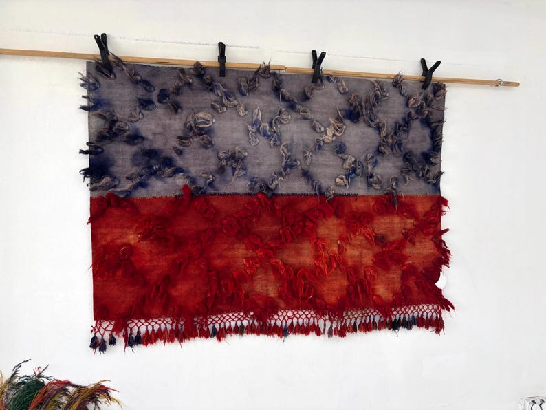 May include: A rectangular textile art piece, divided horizontally into two sections. The top half is a muted blue-gray with textured, raised elements. The bottom half is a vibrant red with a fringed edge, creating a striking contrast.