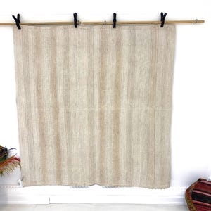 May include: A beige, vertically striped textile hanging from a wooden rod with black clips. The fabric has a woven texture and is displayed against a white wall. The textile is likely a blanket or a curtain.