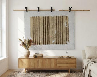 May include: A rectangular wall hanging featuring a light gray fabric base with a centered, distressed brown and tan striped textile panel. The piece is suspended from a wooden rod using four black leather straps, displaying a raw, frayed edge finish.