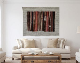 Puede incluir: Un tapiz rectangular con un panel central de rayas verticales en tonos negro, rojo y amarillo, enmarcado por un borde gris texturizado más ancho. La tela central muestra un desgaste visible y bordes deshilachados a lo largo de las costuras.