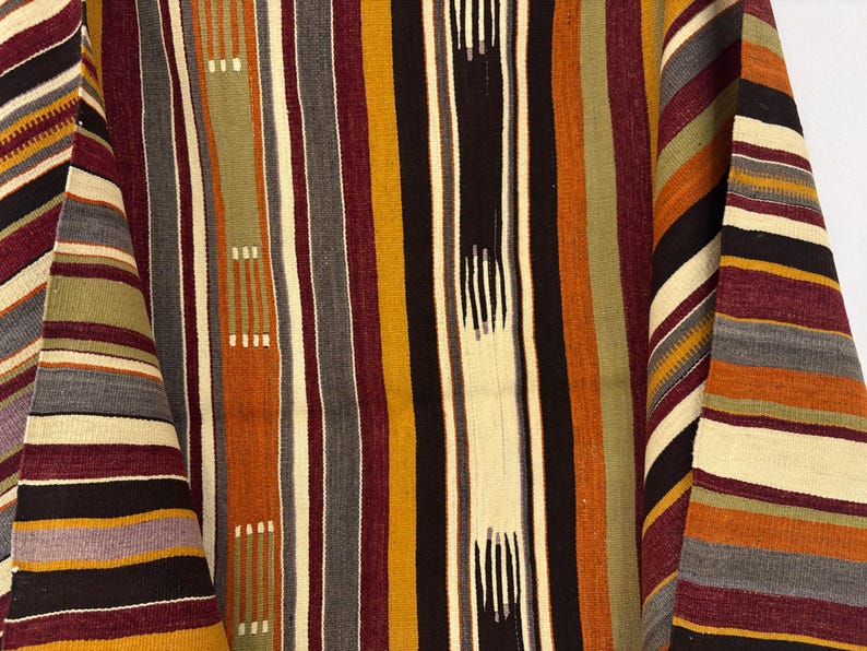 May include: A woven textile featuring a repeating vertical striped pattern in shades of mustard yellow, deep burgundy, olive green, black, cream, and grey. The fabric displays a textured weave with alternating solid and dashed line segments.