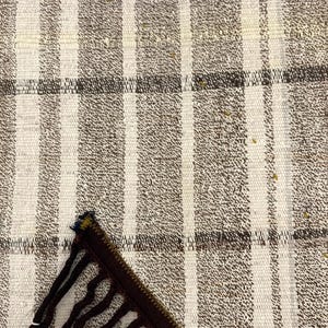 Puede incluir: Primer plano de un textil tejido con un patr&oacute;n de rayas en tonos beige, marr&oacute;n y blanco. La tela tiene un aspecto texturizado, con rayas verticales y una raya horizontal marr&oacute;n m&aacute;s oscura. Se ve un borde con flecos marr&oacute;n oscuro y negro.