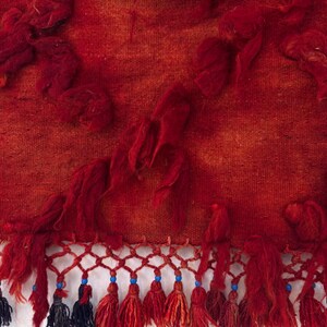 May include: Close-up of a red textile with a textured surface and tufted accents. The bottom edge features a decorative fringe with tassels in red, blue, and gray, adding a handcrafted look.