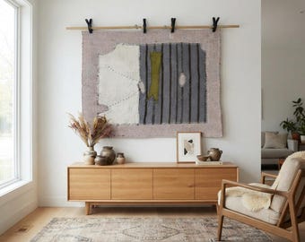 May include: A rectangular wall hanging featuring a light purple base with a white panel and a dark gray striped section, suspended from a wooden rod by four black clips. The textile displays visible stitching and a central olive green fabric accent.