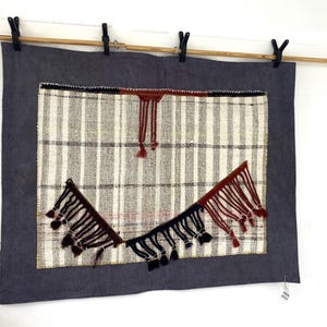 Puede incluir: Tapiz textil de pared con borde gris oscuro y panel central con rayas verticales blancas y grises. Borlas decorativas en negro y burdeos a&ntilde;aden inter&eacute;s visual. La pieza est&aacute; suspendida de una barra de madera con pinzas negras.