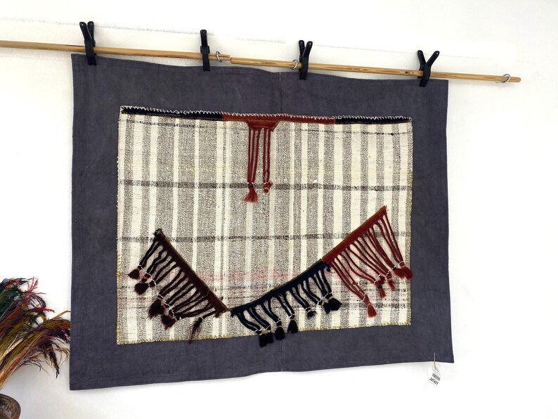 Puede incluir: Tapiz textil de pared con borde gris oscuro y panel central con rayas verticales blancas y grises. Borlas decorativas en negro y burdeos a&ntilde;aden inter&eacute;s visual. La pieza est&aacute; suspendida de una barra de madera con pinzas negras.