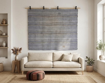 May include: A large, rectangular, woven wall hanging featuring horizontal stripes in shades of blue, gray, and tan. The textile is suspended from a wooden rod with four black clips, displaying a textured surface with subtle variations in color.
