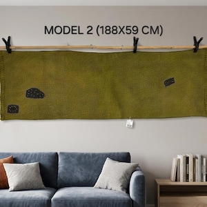 May include: A rectangular olive green textile wall hanging featuring three small black embroidered shapes with white dots. The fabric is suspended from a light-colored wooden rod by three black clips. Text above reads MODEL 2 (188X59 CM).