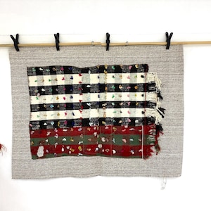 May include: A rectangular textile with a black and white checkered pattern, accented with colorful flecks, is displayed. The textile has a fringed edge and is mounted on a larger gray woven fabric, suspended by black clips on a wooden rod.