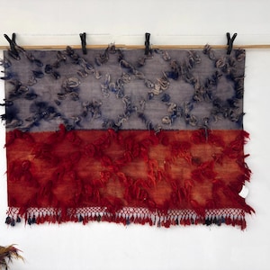 May include: A rectangular textile with a two-tone design. The top half is gray with blue accents, and the bottom half is red with a fringed edge. The textile is suspended from a wooden rod with black clips.
