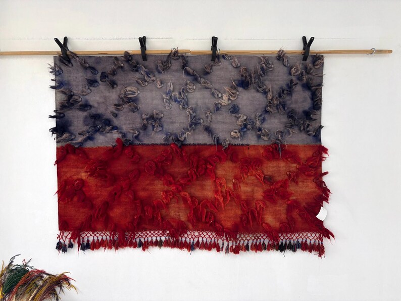 May include: A rectangular textile with a two-tone design. The top half is gray with blue accents, and the bottom half is red with a fringed edge. The textile is suspended from a wooden rod with black clips.