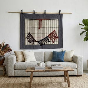 May include: A living room scene featuring a light gray sofa with decorative pillows, a wooden coffee table, and a woven rug. A large wall hanging with a gray border and fringed details hangs above the sofa. A potted plant and dried flowers add natural elements.