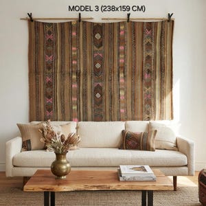 Op de afbeelding: Een groot bruin wandkleed met het label MODEL 3 (238x159 CM) met verticale strepen en geometrische patronen in roze, geel en wit. Het textiel hangt aan een houten roede met zwarte clips boven een cr&egrave;mekleurige bank.