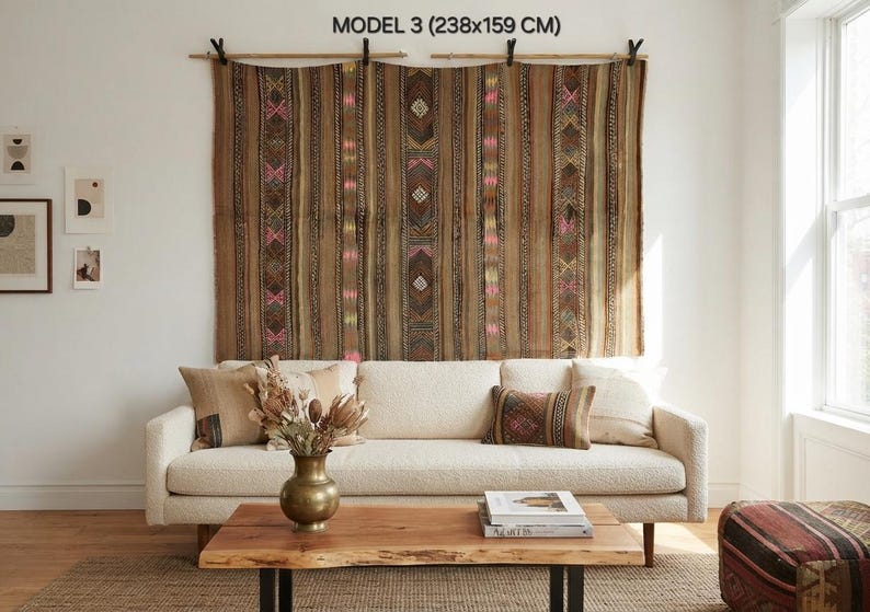 Op de afbeelding: Een groot bruin wandkleed met het label MODEL 3 (238x159 CM) met verticale strepen en geometrische patronen in roze, geel en wit. Het textiel hangt aan een houten roede met zwarte clips boven een cr&egrave;mekleurige bank.