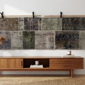 May include: A long, rectangular wall hanging composed of multiple stitched fabric patches in muted shades of olive green, charcoal grey, and brown, suspended from two light-coloured wooden rods by black clips against a plain white wall.