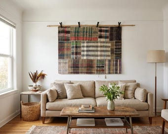 May include: A rectangular wall hanging featuring a patchwork of various plaid and checkered fabric patterns in muted tones of green, brown, blue, and cream, suspended from a wooden rod by four black clips against a plain white wall.