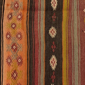 Op de afbeelding: Een geweven textielkleed met verticale strepen in tinten oranje, donkerbruin, olijfgroen en dieprood. De stof toont herhalende geometrische ruitpatronen in cr&egrave;me, rood en geel, ge&iuml;ntegreerd in het gestreepte ontwerp.