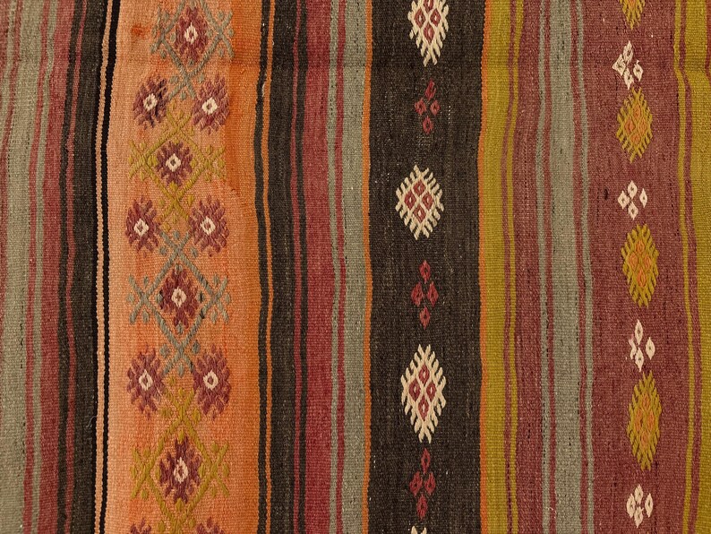 Op de afbeelding: Een geweven textielkleed met verticale strepen in tinten oranje, donkerbruin, olijfgroen en dieprood. De stof toont herhalende geometrische ruitpatronen in cr&egrave;me, rood en geel, ge&iuml;ntegreerd in het gestreepte ontwerp.