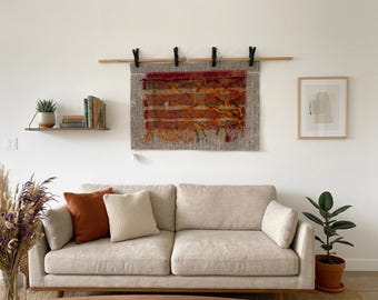 Pode incluir: Uma tapeçaria retangular tecida com listras horizontais em tons de vermelho, laranja e amarelo, suspensa por uma haste de madeira com quatro clipes pretos. A arte têxtil exibe uma aparência texturizada e em camadas com bordas desfiadas.