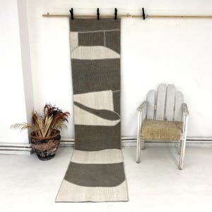 Puede incluir: Una alfombra rectangular larga con un diseño geométrico en tonos grises y blanquecinos cuelga de una barra de madera. El patrón de la alfombra incluye rayas y formas abstractas. Una silla de madera blanca y una planta en maceta también están en la imagen.