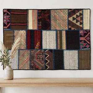 May include: A rectangular wall hanging composed of various woven fabric patches in shades of brown, black, red, cream, and green. The textile features a grid-like arrangement of geometric patterns, stripes, and solid colours stitched together with dark thread.
