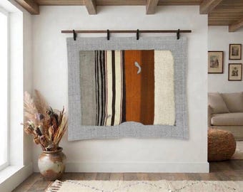 May include: A rectangular wall hanging featuring a central panel of vertical stripes in shades of gray, black, cream, rust orange, and dark brown, framed by a wider light gray border, suspended from a dark wooden rod with four black clips.