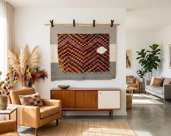 Puede incluir: Un tapiz rectangular con un patrón central de chevrón en negro, rojo y amarillo, bordeado por paneles de tela gris y blanca. El textil está suspendido de una varilla de madera mediante cuatro clips negros sobre una pared blanca lisa.