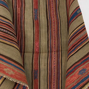 Op de afbeelding: Een geweven textiel met een herhalend patroon van verticale strepen in tinten beige, bruin, rood en blauw. De stof vertoont een getextureerd, grof weefsel met kleine, geborduurde geometrische motieven in blauw en roze die erin verwerkt zijn.