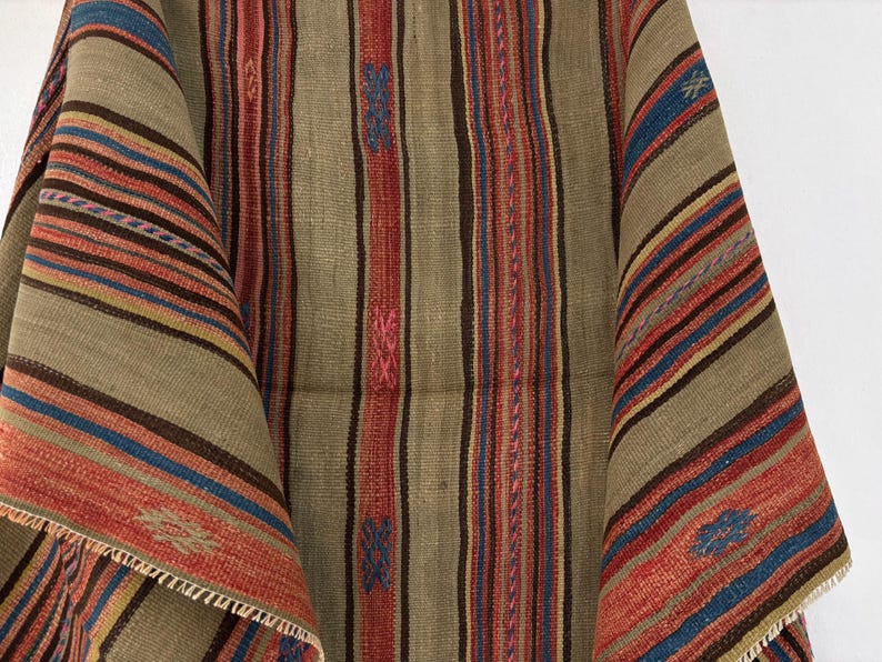 Op de afbeelding: Een geweven textiel met een herhalend patroon van verticale strepen in tinten beige, bruin, rood en blauw. De stof vertoont een getextureerd, grof weefsel met kleine, geborduurde geometrische motieven in blauw en roze die erin verwerkt zijn.