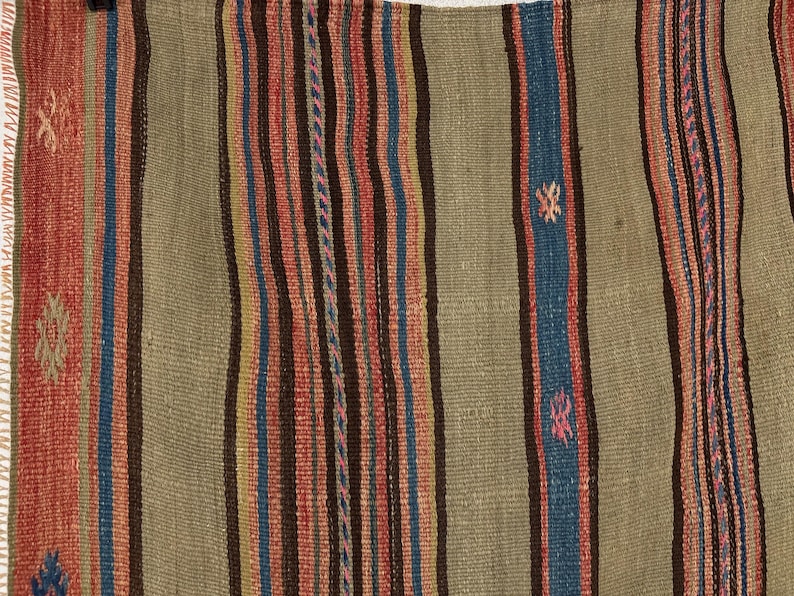 Op de afbeelding: Een geweven textiel met een reeks verticale strepen in tinten olijfgroen, donkerbruin, blauw en rood. De stof vertoont een gestructureerde, platgeweven constructie met kleine, herhalende geometrische motieven ge&iuml;ntegreerd in het streeppatroon.