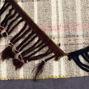 Puede incluir: Primer plano de una secci&oacute;n de textil tejido con ribete de flecos marrones y azul marino. El textil presenta un patr&oacute;n de rayas en tonos neutros con toques de rosa y azul. Los flecos anudados a&ntilde;aden un elemento decorativo a la tela.
