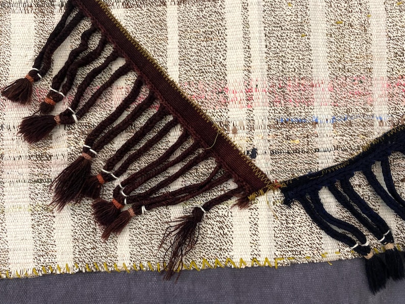 Puede incluir: Primer plano de una secci&oacute;n de textil tejido con ribete de flecos marrones y azul marino. El textil presenta un patr&oacute;n de rayas en tonos neutros con toques de rosa y azul. Los flecos anudados a&ntilde;aden un elemento decorativo a la tela.