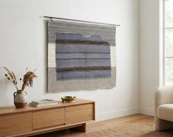 Puede incluir: Un tapiz rectangular tejido que presenta un panel central azul con rayas horizontales de color marrón oscuro, enmarcado por bordes grises texturizados y bandas blancas verticales en los lados, suspendido de una fina varilla de metal oscuro.