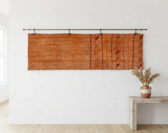May include: A long, rectangular, burnt-orange textile wall hanging suspended from a metal rod by four black S-hooks. The fabric features a textured weave with vertical stripes and small, repeating geometric patterns in darker red tones.