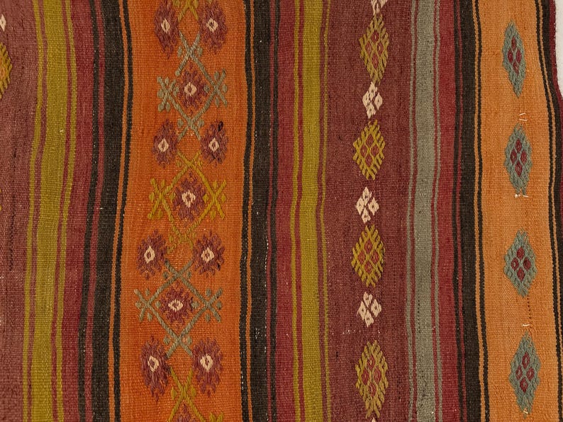 Op de afbeelding: Een geweven textielkleed met verticale strepen in tinten van gebrand oranje, diep kastanjebruin, olijfgroen en donkerbruin. De stof toont herhalende geometrische ruitpatronen in gedempte tinten cr&egrave;me, groenblauw en mosterdgeel over het gehele oppervlak.