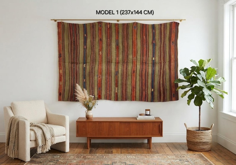Op de afbeelding: Een grote rechthoekige wandhanger met verticale strepen in olijfgroen, roestoranje en donkerblauw, voorzien van kleine geometrische patronen. De tekst MODEL 1 (237x144 CM) staat boven het textiel dat aan een gouden roede hangt.
