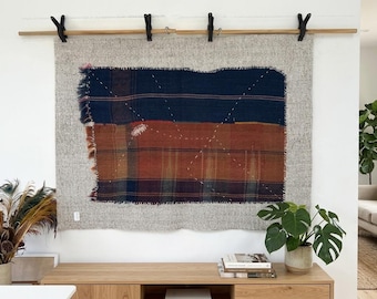 Puede incluir: Tapiz textil de pared con un diseño rectangular en tonos azul marino, óxido y gris. El textil está montado sobre una barra de madera con clips negros. El tapiz se exhibe sobre un mueble de madera.